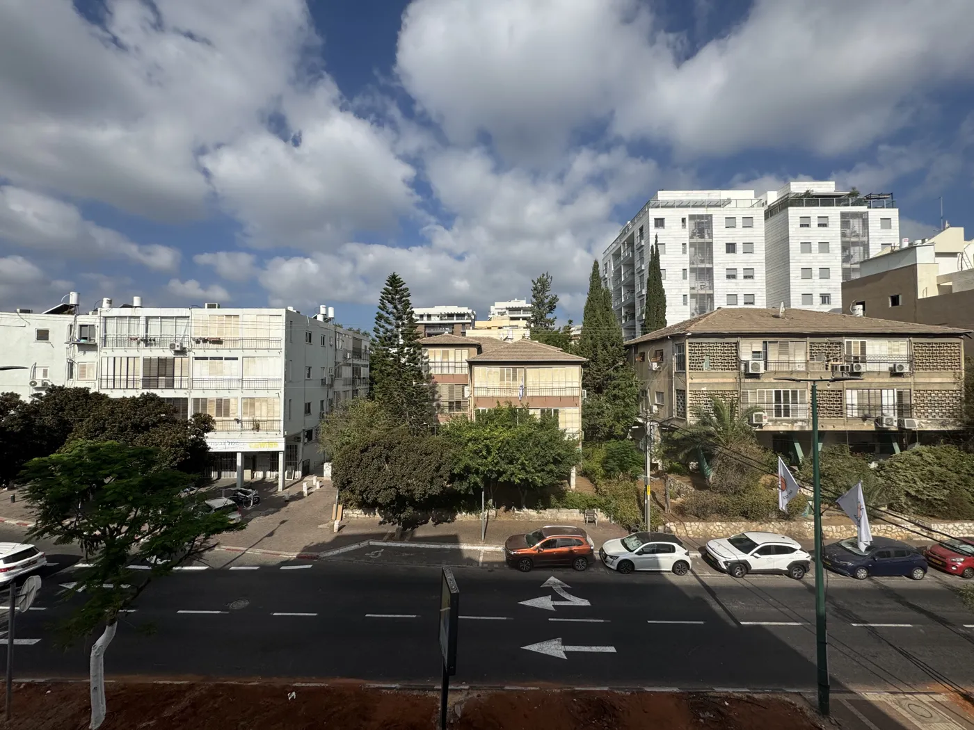 צילום אמיתי מהחלון של רחוב עירוני בישראל: בנייני מגורים בז' ולבנים, שני בתים עם גגות רעפים, ברושים גבוהים, מגדל מגורים לבן מימין, ומכוניות חונות לאורך הכביש עם חצי כיוון על האספלט
